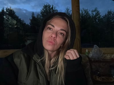 A medium shot captures a woman with blonde hair and brown eyes, wearing a dark hoodie and a green jacket. Her lips are pursed, and she is looking directly at the camera. Her left hand is visible, resting on the sleeve of her jacket. In the background, trees and a cloudy sky are visible, suggesting an outdoor setting. The image is dark, with hints of artificial light. To the right, a wooden post, a patterned couch, and a plastic bag are discernible. The scene is devoid of any discernible urban features, indicating it is likely not in the city of Zheleznodorozhnyi, Russia.