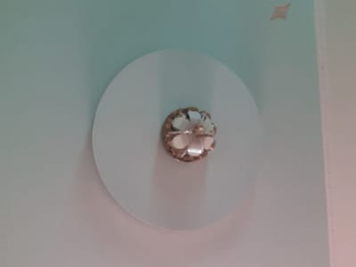 The image shows a close-up view of a decorative element, possibly a light fixture or a ceiling accent, against a pale blue and white background. The central object is a circular, flat white disk, with a smaller, ornate embellishment positioned in its center. This embellishment appears to be made of metallic material and features multiple clear or iridescent petals, resembling a flower. The background is softly lit and textured, with subtle color gradients suggesting a wall or ceiling. There is a small, light brown diamond-shaped mark on the upper right portion of the background. No people or animals are visible. The scene is indoors, likely in a residential or commercial space. The provided image does not contain any specific information that would allow for its location to be identified within the city of Интернационал, Kyrgyzstan.