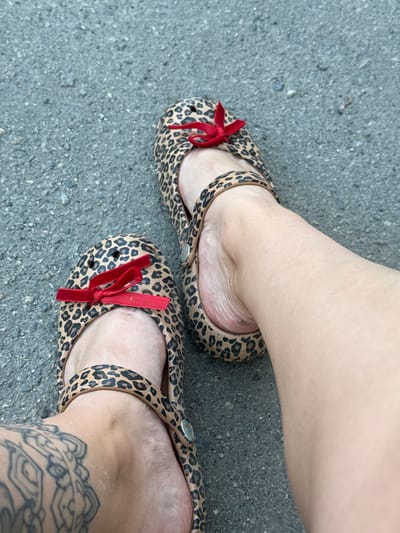 The image shows a close-up, overhead view of a person's feet wearing leopard-print shoes with red bows. The shoes appear to be a type of slip-on or sandal, possibly made of rubber or plastic, with a distinctive pattern of dark brown spots on a tan background. Red velvet bows are tied onto the front of each shoe. One foot is positioned slightly higher and further back than the other, suggesting the person is standing or has just stopped walking. The skin of the feet is smooth and light-toned. A tattoo with intricate, dark linework is visible on the side of the ankle of the lower foot.
The shoes are resting on a gray, asphalt-like surface, which is textured with small stones and pebbles. This surface is consistent with an outdoor setting, such as a sidewalk, pathway, or road. The lighting suggests it is daytime, with no harsh shadows indicating direct sunlight. The overall mood is casual and everyday. The location context provided is Piatykhatky, Ukraine. No text is visible in the image.