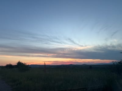 An outdoor landscape shot captures a sunset over a rural area, likely in or around Ivanyane, Bulgaria. The sky transitions from a clear, pale blue at the top to hues of pink, orange, and darker gray clouds near the horizon, indicating the sun has recently set or is about to set. Wispy clouds stretch across the sky. In the foreground, a field of tall, dark green grass and some vegetation stretches across the image, with a dirt path visible on the left. The background features rolling hills and distant, silhouetted structures. The overall lighting suggests twilight. There are no people or animals visible in the frame.