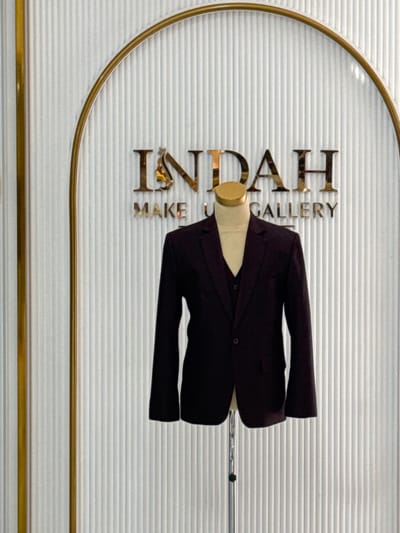 The image shows a mannequin dressed in a black suit with a waistcoat and a jacket, displayed against a vertically striped white wall. Above the mannequin, the word "INDAH" is written in large, metallic gold letters, with the letters "I", "N", and "D" partially visible through a golden archway. Below "INDAH", the words "MAKE U GALLERY" are also in gold lettering. To the left of the display is a vertical gold pole. The overall setting appears to be a store or gallery. The provided context suggests this scene is located in Tanjungmeru, Indonesia.