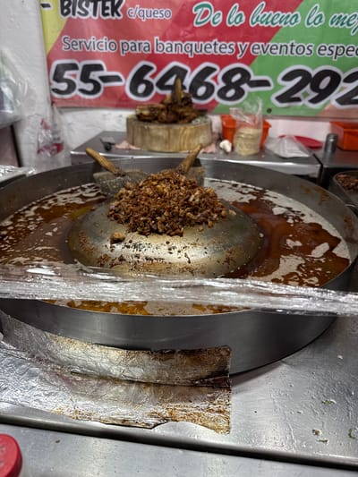 The image shows a close-up of a food preparation area, likely in Tizapan, Mexico. The primary focus is on a large, round metal griddle or cooking surface, splattered with grease and bits of cooked meat. A metal utensil, possibly a spatula, is resting in the center, holding a pile of chopped, seasoned meat. The cooking surface is set within a larger metal apparatus, with a clear plastic wrap draped over part of it, suggesting active food preparation or keeping food warm.
To the left of the cooking surface, a red banner with white and green text is visible. The text, in Spanish, advertises "BISTER c/queso" (Bistek with cheese) and "Servicio para banquetes y eventos espec" (Service for banquets and special events). Prominently displayed are the phone numbers "55-6468-292". The banner's vibrant colors and bold lettering indicate it's an advertisement for a food vendor or catering service.
In the background, glimpses of other food-related items can be seen, including what appears to be a stack of tortillas wrapped in plastic, and a red container. The overall setting suggests a busy, possibly outdoor, food stall or a kitchen preparing food for an event. The lighting suggests it is daytime. The image captures a moment in the process of preparing a common Mexican dish, highlighting the ingredients and the advertising used to attract customers.