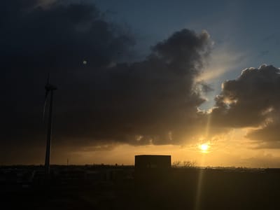 The image displays a landscape in Naaldwijk, Netherlands, during either sunrise or sunset, characterized by a dynamic sky dominated by clouds. The sun is positioned low on the right side of the frame, emitting a bright, warm light through a break in the dark grey and blue-black cloud formations. A prominent vertical lens flare extends downwards from the sun. On the far left, a tall wind turbine stands silhouetted against the dark sky, with its blades angled upwards. In the middle-right, a dark, rectangular building with visible windows is silhouetted against the horizon, near a cluster of bare trees. The foreground and midground are largely obscured by shadow, revealing a flat, indistinct terrain with distant structures and a faint urban skyline, including a communication tower, along the horizon. A small, greenish circular artifact is present in the upper left portion of the sky, and a small, dark bird-like object is visible in the upper central sky.