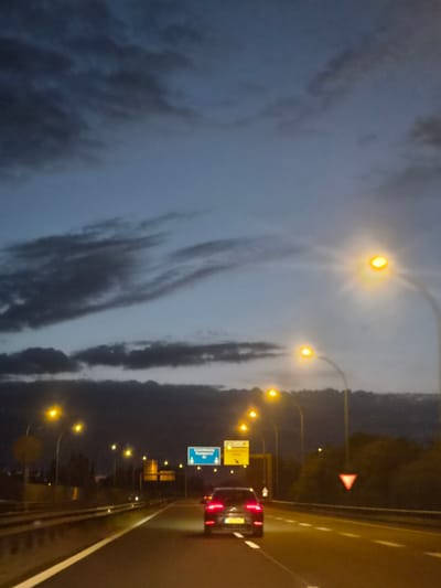 This image depicts a nighttime view from a vehicle on a highway in Mondercange, Luxembourg. The sky is a deep blue with dark, wispy clouds, suggesting dusk or dawn. Numerous streetlights, emitting a bright yellow glow, line the highway, illuminating the road and surrounding trees. A dark-colored car is visible in the foreground, driving ahead on the highway. Traffic signs are present, including a blue directional sign indicating "Luxembourg" and "Pompelle," and a red triangular yield sign. The highway has multiple lanes marked with white lines. The scene is characteristic of a major road in a European country during low light conditions.