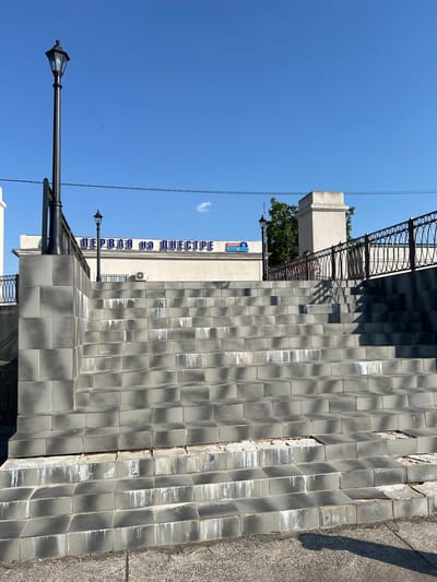 The image depicts a wide outdoor staircase made of grey tiles leading up to a building. The building has blue Cyrillic lettering that reads "ПЕРВАЯ на ДНЕСТРЕ", which translates to "First on the Dniester". A lamppost stands to the left of the stairs, and a black metal railing runs along the side. The sky above is clear and blue. The scene appears to be located in Korzhevo, Moldova. The condition of the staircase shows some wear and tear, with missing and damaged tiles in places, and white streaks running down some of the steps, possibly from water damage. There are no people visible in the image.
