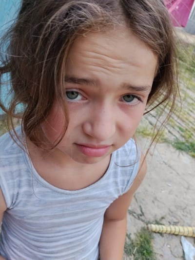 A close-up, high-angle shot focuses on the face of a young girl with brown hair and blue-green eyes. She is wearing a light gray, horizontally striped tank top. Her forehead shows visible wrinkles, and her expression appears somber or concerned. Her hair is disheveled and falls around her face.
The background is outdoors and appears to be a rural or somewhat neglected area. To the left, a light blue wall is visible, and to the right, a textured ground surface that looks like a pathway or pavement, interspersed with patches of green grass. A yellow object with green wrapping, resembling a rolled-up mat or similar item, lies on the ground in the lower right corner.
The lighting suggests it is daytime, possibly late afternoon given the angle of light and shadows, although this is not definitively clear. The overall mood conveyed by the girl's expression and the slightly unkempt setting is one of seriousness or perhaps a moment of sadness or contemplation. There is no visible text in the image.
