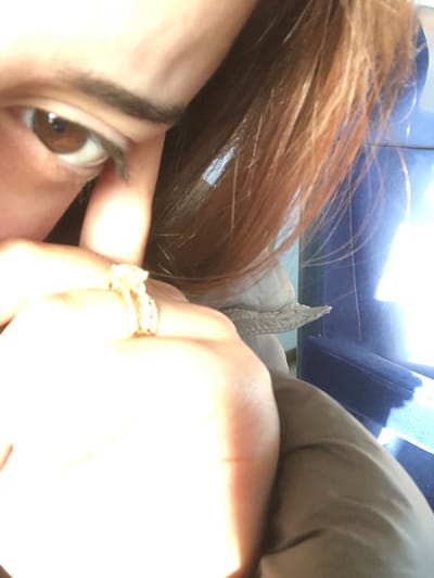 This image provides a close-up, highly illuminated view of a person indoors in Dettwiller, France. The upper left quadrant reveals a brown eye with dark eyelashes and eyebrow. Brown hair frames the right side of the face. In the immediate foreground, a hand, heavily overexposed by a bright light source, displays two rings on one finger: an engagement-style ring featuring a prominent clear central stone surrounded by smaller stones on a gold band, and a second plain gold band. The person is wearing a tan or brown sleeve, with a lighter, possibly off-white, textured fabric visible behind the hand. In the background, a segment of a deep blue upholstered armchair is discernible.