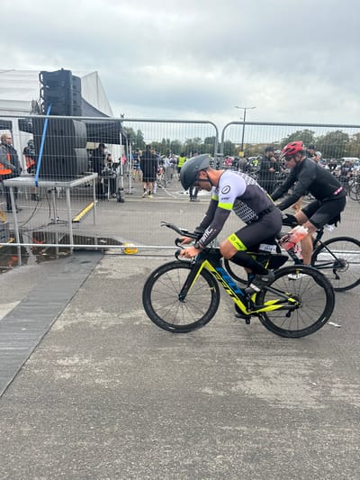 The image displays an outdoor event scene in Weymouth, United Kingdom, characterized by an asphalt surface and an overcast sky.
In the immediate foreground, two male cyclists are positioned on their bicycles. The first cyclist, on the left, wears a black, white, and yellow long-sleeved aerodynamic cycling suit, a black helmet, and glasses. He is on a black and yellow Felt time trial bicycle with disc wheels, leaning forward with hands on the aerobars. A race number "2018" is affixed to his seat post.
To his right, the second cyclist wears a black long-sleeved top, black cycling shorts, a red helmet, and sunglasses. He is seated on a black road bicycle, which has a bottle containing an orange liquid in a frame-mounted cage. A race number "2021" is visible on his seat post.
The midground and background feature event infrastructure. On the far left, multiple black sound system speakers are stacked on metal scaffolding, positioned near a large white event tent. Metal crowd barriers run across the scene, separating the immediate area from a gathering of numerous individuals and additional bicycles further back. Some individuals in the background wear high-visibility vests. A dark grey event mat is laid across the asphalt surface in the left mid-ground, adjacent to a visible puddle of water. Distant trees and a street lamp pole are visible against the cloudy sky.