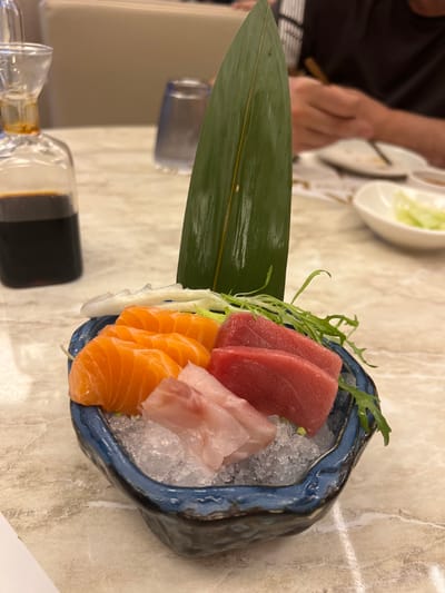 A plate of assorted sashimi is presented on a table with a marbled surface. The sashimi includes slices of salmon, tuna, and white fish arranged on a bed of crushed ice within a dark-colored bowl with a blue rim. A large green leaf is positioned to the left of the bowl, acting as a garnish. A small container filled with soy sauce is also visible on the left side of the frame. In the background, a person is seen holding chopsticks, and a shallow bowl with a light-colored salad is partially visible. The setting appears to be a restaurant. The location, Torreforta, Spain, cannot be determined from this image alone.