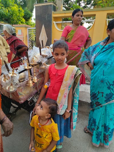The image, taken in Kammavaripalli, India, shows a group of people on a paved outdoor surface near a building. A young girl in a red shirt and blue skirt stands in the foreground, with a patterned cloth draped over her shoulder. A small child in a yellow shirt with a mustache print stands next to her, looking upwards. Behind them is a cart laden with brass oil lamps, many still wrapped in plastic. To the right of the girl, two women are visible, wearing traditional saris. The first one is wearing a pink and green sari and the second one is wearing a blue sari with floral patterns. To the left of the cart, an older woman in a red and black patterned sari stands near a yellow and white gate. The background includes a building with a gray and yellow facade and trees. The day's lighting is bright and casts shadows, suggesting it's daytime.