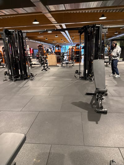 The image depicts the interior of a fitness gym located in Aubange, Belgium. The floor is covered with dark grey interlocking rubber mats. Overhead, exposed wooden beams support a ceiling with suspended dome-shaped lights, silver-colored ventilation ducts, and electrical conduits.
In the foreground, a black adjustable utility bench is positioned towards the right, casting a shadow to its left. To the left of the frame, a large black cable machine with multiple pulleys and weight stacks is visible. Another similar cable machine stands in the mid-ground to the right.
One male individual stands in the mid-ground right, facing left. He is wearing a grey hooded sweatshirt with dark stripes on the sleeves, dark trousers, and white athletic shoes. Over-ear headphones are on his head, and he is looking down at a mobile phone held in both hands.
In the background, numerous pieces of orange and black gym equipment are arranged, including various weight machines and benches. A light-colored wooden cabinet or counter is visible in the mid-background. Several other individuals are present in the background, utilizing or standing near the gym equipment. One person is observed lying on a bench in the mid-background left.