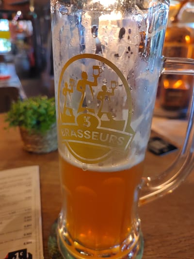 The image shows a close-up of a tall glass of amber-colored beer with a foamy head. The glass has a logo with silhouettes of three people holding beer mugs and the text "3 BRASSEURS". The glass is condensation-covered and sits on a wooden table. In the background, out of focus, a small green potted plant is visible to the left, and a blurry image of a building interior with lights can be seen further back. A menu or bill is partially visible on the left side of the image, showing some text and prices. The overall impression is that of a casual dining or drinking establishment. This scene could be located at the "3 Brasseurs" restaurant in Éragny-sur-Oise, France, as indicated by the brand name and logo visible on the glass.