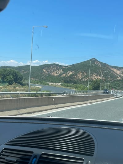The image presents an eye-level view from inside a moving vehicle on a multi-lane highway, looking forward towards a mountainous landscape, specifically within the Δημοτική Ενότητα Προμαχώνος region of Greece. The lower portion of the frame shows the dark grey dashboard of the vehicle, featuring prominent horizontal and semicircular air vents.
The highway occupies the mid-ground and extends into the foreground, with clear white lane markings on the asphalt. A concrete median barrier separates traffic lanes, and a metal guardrail runs along the edge of the bridge. The road traverses a bridge over a dark, calm body of water, likely a river, which winds through the landscape below the bridge. Sparse, dry-looking vegetation and some denser green trees line the riverbanks.
Beyond the river, the background is dominated by a range of green, tree-covered mountains. Distinct patches of lighter brown, exposed earth or rock formations are visible on the mountain slopes, suggesting geological features or areas with less vegetation. One prominent mountain peak rises in the center-right background.
Several tall, grey streetlights with single luminaires are positioned along the highway, extending over the road. Power lines are faintly visible horizontally across the mid-ground against the mountains. Ahead on the bridge, a dark SUV is discernible, followed by a white sedan, and other vehicles are present further in the distance. The sky above is clear and bright blue with a few scattered white cumulus clouds near the horizon. Small smudges or debris are visible on the vehicle's windshield, indicating the perspective is through glass. The scene is illuminated by bright daylight.