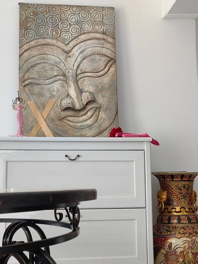 The image shows a close-up of a room's interior, likely a living area or hallway, in Квант-88 Снт, Russia. The focal point is a large, decorative wall piece depicting a Buddha's face, intricately carved with swirling patterns. To the left of the Buddha, on top of a white cabinet with a single drawer, sits a pink, mannequin-like jewelry holder and a wooden "X" sculpture. A red rose and some pink fabric are also placed on the cabinet. To the right of the cabinet, a large, ornate vase with golden elephant heads and intricate patterns stands prominently. In the foreground, a dark, round table with a decorative metal base is partially visible. The lighting suggests an indoor environment with natural light entering from the side.