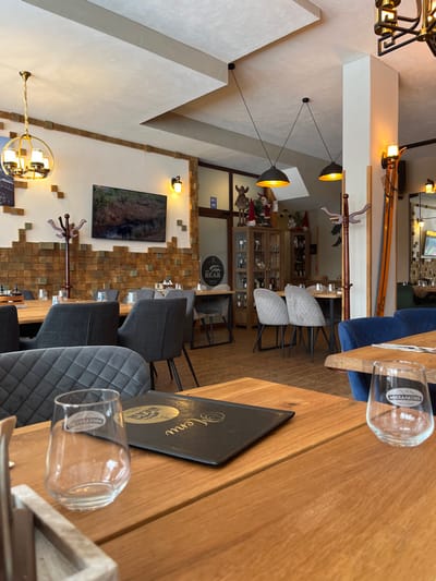 The scene captures the interior of an unoccupied restaurant in Stoikite, Bulgaria. In the immediate foreground, a light-colored wooden table features a black menu folder with gold lettering and a clear, empty drinking glass bearing the "МИХАЛКОВО" brand and a mountain logo. A dark grey quilted chair is positioned to the left. The midground reveals a dining area with multiple light-colored wooden tables and a mix of dark grey and lighter grey quilted upholstered chairs. No individuals are present. The left wall displays a patterned design of light brown wooden blocks, above which a flat-screen television shows a natural landscape. A golden wall sconce and a brown wooden coat rack are on this wall. A golden, multi-tiered chandelier hangs from the ceiling on the left, and two black pendant lamps with golden interiors hang centrally. Towards the rear, a doorway with a "WC" sign leads to another section. Adjacent to this, a tall wooden display cabinet with glass doors is stocked with bottles, topped with decorative figures including a large moose, Santa Claus figures, and gnomes. A circular "BEAR" logo sign is mounted on the wall nearby. On the far right, a pair of vintage-style wooden skis leans against a wall, illuminated by another golden wall sconce. A mirror on the right reflects additional seating. The ceiling features a multi-level design in white and light grey tones.