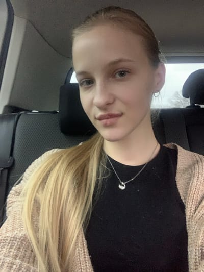 A young fair-skinned female with light blonde hair, pulled back into a ponytail, is depicted in a close-up, chest-up view, seated inside a vehicle in Oliivka, Ukraine. She has light-colored eyes and full lips, with small hoop earrings visible. She is wearing a black top beneath a light beige or brown knitted cardigan and a silver-colored necklace with a small, curved pendant. Her gaze is directed towards the camera. The background shows the dark interior of the vehicle, including parts of the rear seat and headrests. A window in the upper right reveals a blurred, overcast sky or outdoor scene.