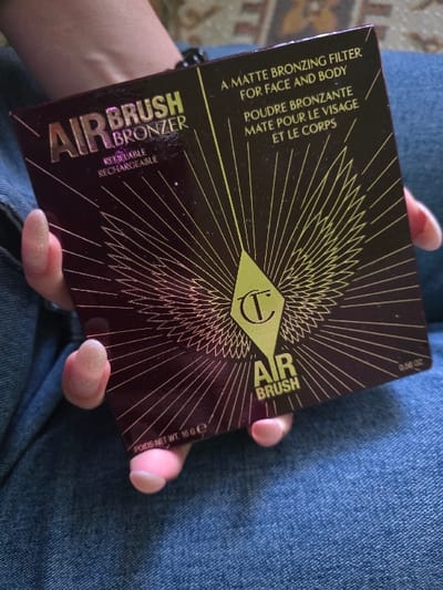 A human hand, featuring long, oval-shaped nails with light pink glitter polish, holds a rectangular Charlotte Tilbury "AIRBRUSH BRONZER" product box. The box is dark burgundy with gold text and a central stylized "CT" logo surrounded by radiating lines. Visible text includes "REFILLABLE RECHARGEABLE," "A MATTE BRONZING FILTER FOR FACE AND BODY," "POUDRE BRONZANTE MATE POUR LE VISAGE ET LE CORPS," "POIDS NET WT. 16 G E," and "0.56 OZ." A black bracelet is visible on the wrist. The hand is positioned against a background of blue denim fabric, likely jeans worn by the individual, with an out-of-focus, patterned textile visible in the upper background, suggesting an indoor setting. No visual information in the image indicates the location as Staliyska mahala, Bulgaria.