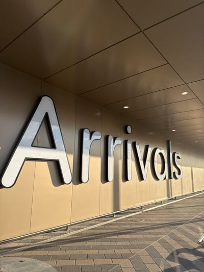 The image depicts a building facade with the name "Airwals" displayed vertically in large, white letters with black outlines. The letters are attached to a smooth, beige-colored wall composed of rectangular panels. Small, circular lights are embedded in the wall above the lettering. A brick wall with a metallic handrail is visible on the right side of the frame. The angle suggests the photograph was taken from below, looking upwards at a slant. This is located in Crumlin, United Kingdom.