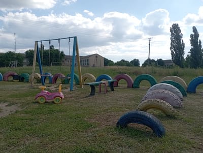 The image depicts an outdoor playground area, likely located in Inhulo-Kamianka, Ukraine. The foreground is dominated by a grassy area with a dirt path running through it. In the center left of the image, a pink toy car with yellow accents sits on the grass. Behind the toy car, a swing set with blue and yellow supports and chains is visible. The midground features a collection of colorful, repurposed tires arranged in various configurations. Some tires are upright, forming an enclosure, while others are semi-circular and partially embedded in the ground, creating a decorative border. A low bench made of wood and metal supports stands amongst the tires. The background reveals a row of green trees and shrubs, behind which several buildings are situated. One building appears to be a light-colored, multi-story structure, possibly a school or community center. Further back, a cell tower and utility poles with power lines are visible against a sky with scattered clouds. The overall scene suggests a children's play area that has been creatively outfitted with recycled materials in a rural or semi-rural setting.