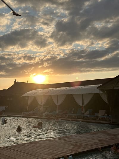 An outdoor swimming pool area is depicted at sunset, with the sun prominently visible in the sky behind a building's rooftop. The sky is filled with dramatic clouds, tinged with the warm hues of the setting sun. In the foreground, a wooden deck leads to the pool, where several individuals are swimming and relaxing. The pool area is furnished with lounge chairs and covered cabanas. A lamppost is visible in the upper left corner of the image, extending from off-frame. The scene is set in Yaroslavskaya, Russia.