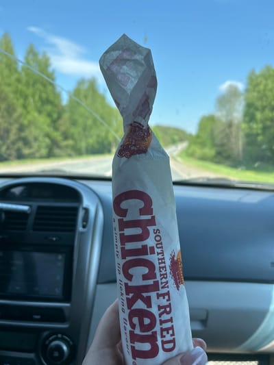 A person is holding a wrapped Southern Fried Chicken sandwich in a car. The interior of the car is visible, including the dashboard, vents, and a screen. Outside the car window, there is a view of trees and the sky. The location cannot be determined from the image.