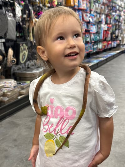 A young child, likely a toddler or preschooler with light hair, is captured smiling brightly and looking upwards in an indoor setting, possibly a shop or market in Ustroń, Poland. The child wears a white t-shirt with ruffled sleeves, featuring pink text that appears to say "Bella" and a graphic of a yellow lemon with green leaves. A brown and green toy snake is draped around their neck, with its head visible near the child's right shoulder. The background is blurred but shows shelves stocked with various items and colorful packaging, typical of a retail environment. The child's joyful expression and the playful toy suggest a moment of happiness and curiosity.