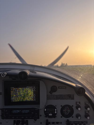 The image depicts a forensic view from inside the cockpit of a light aircraft at Toussus-le-Noble Airport (LFPN), France, during either sunrise or sunset. The lower half of the frame shows the aircraft's instrument panel. On the left, a rectangular digital display, likely a GPS or MFD, shows a detailed map with roads and green areas, along with a digital time of "17:53". Below this display, a Garmin radio unit displays "120.750" as the active frequency and "123.500" as the standby frequency. To the right of these instruments, various analog gauges, knobs, and switches are visible, including a circular gauge with a needle pointing to approximately "20". A white placard on the panel contains regulatory text in English regarding EASA CS-LSA VFR-DAY/NIGHT operations, prohibiting flights in known icing conditions, aerobatic maneuvers, and spins, referencing the airplane flight manual for further limitations.

Through the aircraft's windshield, which exhibits minor smudges and reflections, the external environment is visible. Two vertical stabilizers (tails) of other aircraft are present in the middle distance, out of focus; the left tail has a distinctive red tip. Behind these aircraft, a flat, dark surface consistent with an airfield runway or taxiway is discernible, with a line of trees or low structures on the distant horizon. The sky transitions from a pale blue at the top to a vibrant orange-yellow glow on the right, indicating the low position of the sun. No people are visible within the frame. The aircraft appears to be stationary or taxiing on the ground.