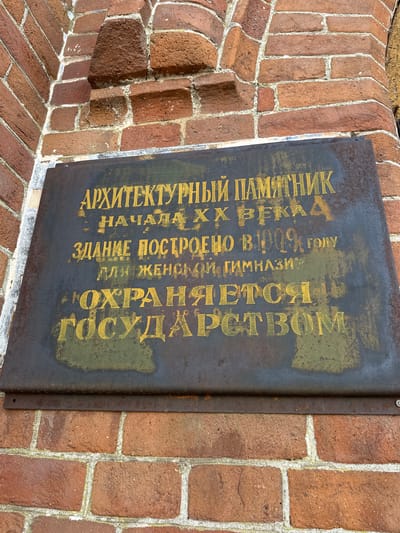 An exterior, eye-level, medium shot shows a weathered, dark metal plaque affixed to a red brick wall. The plaque is inscribed with yellow text in Russian, indicating it is an architectural monument. The text reads: "АРХИТЕКТУРНЫЙ ПАМЯТНИК НАЧАЛА ХХ ВЕКА ЗДАНИЕ ПОСТРОЕНО В 1909 ГОЛУ ДЛЯ ЖЕНСКОЙ ГИМНАЗИИ". Below this, a second line reads: "ОХРАНЯЕТСЯ ГОСУДАРСТВОМ". The plaque is slightly rusted and the text is faded in places. The brick wall shows signs of age and weathering. The image is taken in the city of Nogornyi, Russia.