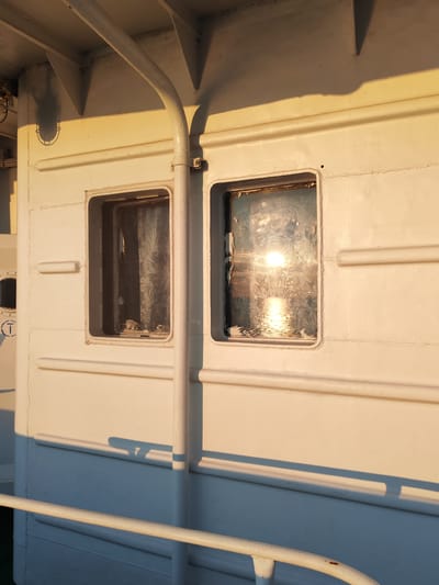 The image shows the side of a white vessel, likely a ship or ferry, bathed in the warm, low light of the setting sun. Two square windows are visible on the hull. The left window is partially obscured by a patterned curtain, while the right window reflects the bright sun, creating a glare that obscures the view within. The hull is made of riveted metal panels and features a horizontal railing in the foreground. Shadows cast by the vessel's structure, including a pipe and an overhead structure, stretch across the hull and the deck below. The environment appears to be a port or waterfront, with the sun setting over a body of water. There are no people or animals visible in the image. The vessel's white paint is slightly weathered. No specific details suggesting the location is Kakkarovo, Russia are present in the image itself.