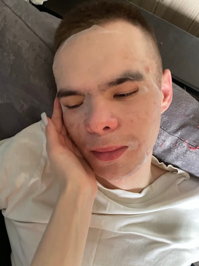 The image shows a person lying on a grey cushioned surface. The person has a facial mask on, covering most of their face. The person's eyes are closed. Their hair is short. One hand is resting on their cheek. They are wearing a white t-shirt. The background is blurred. There is no indication of the location being in Ulukulevo, Russia.