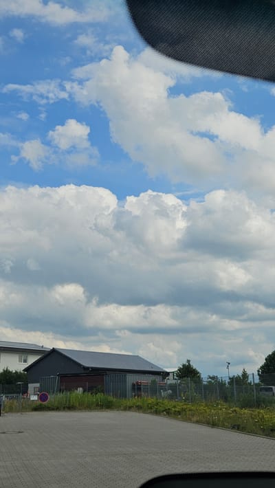 The image depicts an outdoor scene in Buchholz in der Nordheide, Germany. The sky is filled with large, fluffy white cumulus clouds against a bright blue background. In the middle ground, a dark-colored building with a grey roof is visible, flanked by shipping containers. In front of the building, tall green grass and yellow wildflowers grow. A paved area, likely a parking lot, occupies the foreground. A circular red and blue sign is present on the left side of the paved area. The scene appears to be a commercial or industrial area with some natural elements.