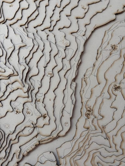 The image shows a topographical map model, likely made of laser-cut wood or cardboard. The model depicts a rugged terrain with contour lines indicating elevation changes. Scattered across the surface are small, abstract representations of buildings or structures, some clustered together to suggest a village or settlement. Faint lines, possibly indicating roads or paths, are also visible. The overall aesthetic is neutral and monochromatic, focusing on form and texture. The question about the location in Piacenza, Italy, cannot be answered from the visual information provided, as the image does not contain any identifying landmarks or textual clues related to a specific place.