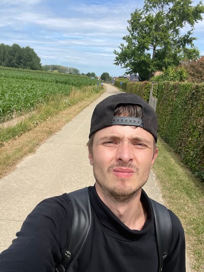 A man wearing a black baseball cap and black long-sleeved shirt is taking a selfie. He is standing on a paved path with a field of green crops on his left and a hedge and houses on his right. The path appears to be in a rural area, possibly on the outskirts of Diepenbeek, Belgium. The sky is blue with scattered clouds. The man's facial expression is ambiguous.