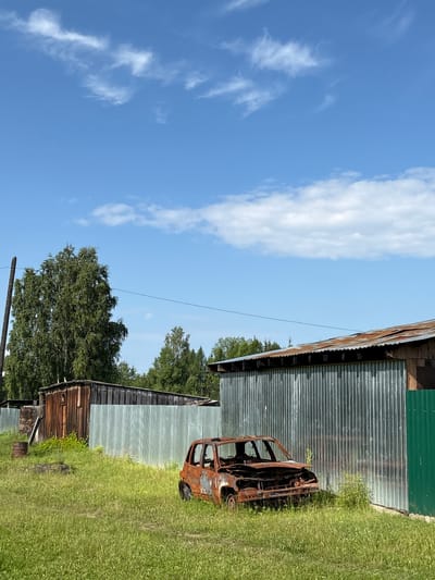 A severely rusted and burned-out car sits in a grassy field in front of corrugated metal buildings. The car, a hatchback, is missing its windshield, hood, and most of its interior. Its wheels are still attached, but the tires appear to be degraded. A rusty barrel stands to the left of the car. Behind the car, a long, weathered wooden building stands next to a section of corrugated metal fencing. Further back, a larger corrugated metal building with a rusted roof is visible. A tall, leafy tree is situated behind the wooden building. The sky above is blue with scattered white clouds. The scene appears to be located in an undeveloped or rural area, possibly on the outskirts of the city of Badarminsk, Russia. There are no people or animals present in the image.