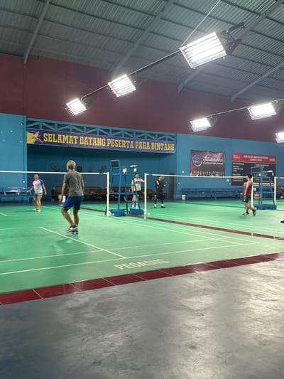 The image captures an indoor badminton court scene, likely during an evening or night session given the prominent stadium lighting. The setting is a sports hall with a green-surfaced court marked with white lines, and the surrounding floor is concrete.
Several individuals are present on the court, engaged in what appears to be a badminton game or practice. In the foreground, a man in a dark grey t-shirt and blue shorts is standing on the court. Further down, another player in a white top and dark pants is near the net. On the opposite side of the net, a player in a maroon top and dark shorts is positioned near the backline, while another player in dark attire is closer to the net. A white shuttlecock is visible in the air near the net.
The ceiling features a corrugated metal roof structure with numerous bright rectangular stadium lights hanging down, illuminating the court. The back wall of the court is blue, transitioning to maroon higher up. A large blue banner stretches across the wall with the Indonesian text "SELAMAT DATANG PESERTA PARA BINTANG" (Welcome Participants, Stars). Above this, a smaller banner displays "Silaturahmi" (Friendship/Gathering) and the acronym "PGRI" (Indonesian Teachers Association), suggesting an event related to teachers' gathering. A red poster on the wall is partially visible, showing text like "CALL OUT FOR CHAMPION LEAGUE." The word "PEGASUS" is prominently written in white on the green court surface. The overall scene depicts an active sports facility in Kecamatan Cisaat, Indonesia.