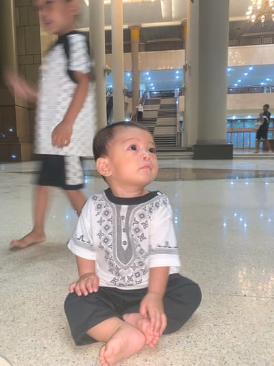 A young child is captured sitting on a light-colored, speckled, and highly reflective floor within a large indoor setting. The child, appearing to be a toddler, is barefoot and wears a white short-sleeved shirt adorned with black and grey geometric patterns, paired with plain black pants. They are positioned towards the lower-right of the frame, looking upwards and slightly to the right with a calm and curious expression, their hands resting on their feet.

The polished floor creates a prominent reflection, displaying a blurry, elongated figure of another child (possibly the same child or another) to the left of the main subject. The background features substantial light grey pillars, suggesting a multi-level architectural space with visible railings and what might be stairs or balconies in the distance. Further back, indistinct figures of adults can be faintly seen. The interior is brightly lit by artificial lights, which cast star-like reflections across the shiny floor surface. The setting appears to be a modern and spacious public building, such as an atrium or a large hall, consistent with urban environments in Samarinda, Indonesia. There is no visible text in the image.