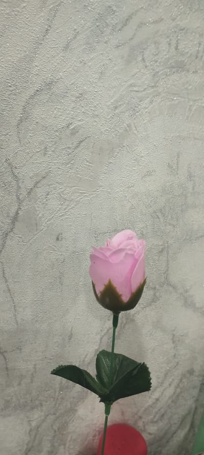 A close-up, vertical, low-angle shot depicts a single, artificial pink rose against a textured, light gray wall with subtle dark gray marbling. The rose is positioned in the lower-middle section of the frame, with its stem extending upwards. The rosebud is closed, revealing layers of pink petals, and a dark green calyx encircles its base. Two green artificial leaves, with a ribbed texture, are attached to the stem below the bud. The background wall has a rough, stucco-like finish with faint, wavy lines of a darker gray. The lower portion of the image is out of focus and shows a blurred red object, possibly a vase or decorative item, from which the rose stem emerges. The lighting is soft, creating a gentle shadow from the rose onto the wall. The overall scene is static and appears to be indoors. The location of Покосное, Russia, is not discernible from the visual information provided in the image.