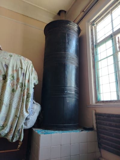A tall, black, cylindrical metal stove stands on a tiled base in a room. The stove has horizontal ribbed bands around its circumference. To the left of the stove, a draped light green fabric with a floral pattern hangs over a piece of furniture. A window with a metal grate is visible to the right of the stove. A dark, ribbed radiator is mounted on the wall beneath the window. The walls are a light peach color, and the ceiling is white. The scene is located indoors.