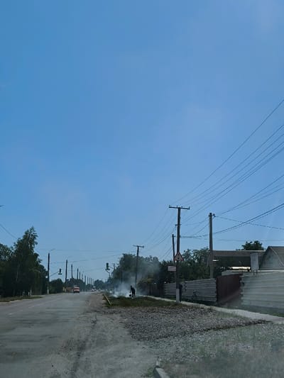A clear blue sky dominates this daytime exterior image captured in Illintsi, Ukraine. The scene depicts a two-lane asphalt road with a worn surface and gravel shoulders extending into the distance. On the right shoulder, a lone individual, dressed in dark attire, operates a device that generates a substantial plume of white smoke. Adjacent to this activity, a large deposit of grey gravel covers part of the roadside. To the immediate right, a corrugated metal fence borders partially visible residential structures. A bicycle is parked against this fence. Numerous wooden utility poles with multiple overhead power lines are positioned along both sides of the road. A triangular warning sign and a rectangular white sign with the Cyrillic text "ОБЕРЕЖНО, ВИСОКА НАПРУГА" (CAUTION, HIGH VOLTAGE) are affixed to a utility pole on the right. Green trees and other vegetation line both road margins. In the mid-distance on the road, a light-colored truck and additional vehicles are visible, alongside traffic signals.