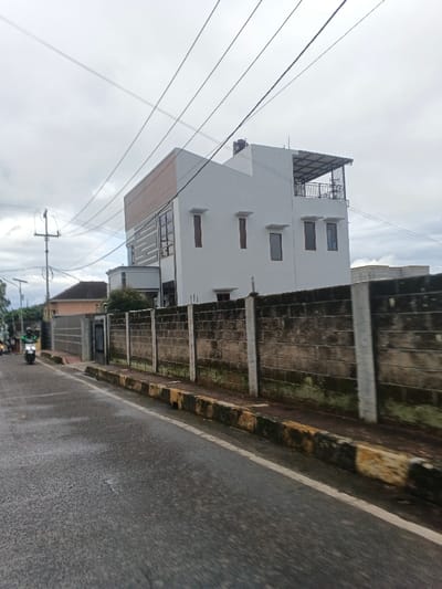 The image depicts a street scene in the city of Masaiti, Zambia, under an overcast sky. A wet asphalt road occupies the foreground, bordered on the right by a concrete curb painted with faded alternating yellow and black stripes. Beyond the curb, a weathered perimeter wall constructed of grey concrete blocks with vertical white concrete posts extends horizontally across the midground. The wall exhibits patches of green and dark biological growth.
Behind the wall, a multi-story white building with a modern architectural style is visible. This building features various rectangular windows with dark frames, some appearing reddish-brown. The upper section on the left includes grey textured horizontal panels. On its rooftop, a dark water tank is present, along with a structure featuring a dark metal frame and a covering, extending over a small balcony with a dark railing on the upper right side. Further down the street on the left, other residential structures, including one with a reddish-brown roof, are partially visible.
In the left foreground, a dark-colored motorcycle or scooter is in motion, heading away from the viewer. The rider is wearing a bright green long-sleeved garment and a dark helmet. A utility pole with crossarms and multiple overhead power lines strung against the grey sky is located on the left side of the road.