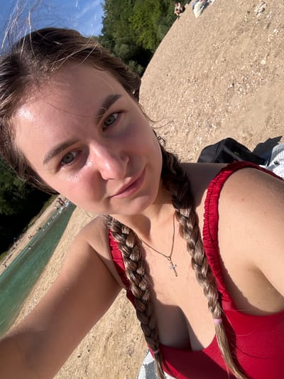 A young woman with blonde hair in two braids, wearing a red bathing suit, is taking a selfie on a sandy beach next to a body of water. She is in the foreground, and the background shows trees, a blue sky with some clouds, and other people in the distance enjoying the water and the beach. The location is the lake in Maisod, France.