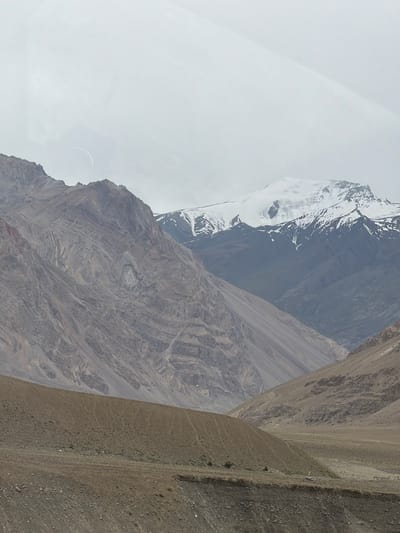 This is a scenic, eye-level, outdoor, vertical, full shot of a mountainous landscape in Zangla, India, under an overcast sky. In the distance, snow-capped mountains rise, with a bright, curved object, possibly a reflection or an artifact, visible in the sky above the left mountain range. The foreground shows a barren, brown, dirt slope with some sparse vegetation. The middle ground is dominated by rugged, layered mountains in shades of brown and gray, with visible striations suggesting geological formations. The background features more distant mountains, some with patches of snow. The overall scene is one of a vast, arid, and remote mountain environment.