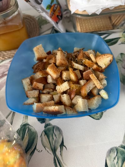 The image shows a close-up, overhead view of a bright blue bowl filled with homemade croutons. The croutons are cut into various irregular shapes and sizes, with some pieces appearing golden brown and toasted, while others are lighter in color, indicating varying degrees of doneness. Some croutons have visible herbs or spices sprinkled on them. The bowl is placed on a tablecloth with a floral or botanical pattern featuring large green leaves and possibly bird motifs in shades of green, white, and muted colors. In the background, to the left of the bowl, is a glass jar with a metallic lid, containing a viscous golden liquid, likely honey or syrup. Partially visible behind the bowl, on the right, is a clear glass container with a lid, filled with square-shaped crackers or biscuits. A white paper napkin or paper towel is folded and placed on top of this container. To the far left, a portion of a clear plastic bag containing a yellowish-orange food item is visible. The scene appears to be set indoors on a table. There is no indication of the city of Crokhovo, Russia, in the image itself.