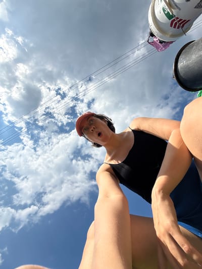 The image shows a low-angle, wide-shot perspective of a woman with a pink baseball cap and glasses looking upwards with a surprised expression. She is wearing a black tank top and blue shorts. Her arms are visible, with one hand resting on her leg and the other reaching towards the camera. In the background, there is a blue sky with white clouds, and power lines stretch across the frame. Two buckets, one white and one black, are visible hanging from the power lines. The white bucket has some red and green markings and a pink plastic bag is tied to it. The scene appears to be outdoors, possibly in an urban or suburban area, given the presence of power lines and buckets. There is no information in the image to indicate the specific location in the city of Dokshitsy, Russia.
