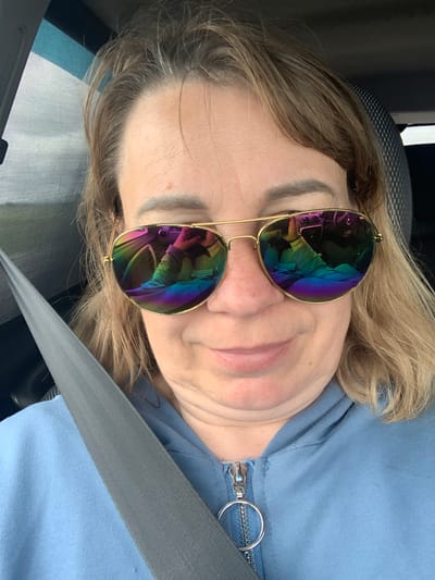 A woman with blonde hair and sunglasses is taking a selfie in a car. The sunglasses have rainbow-colored lenses that reflect the car's interior, including the dashboard and a person's legs. The woman is wearing a blue hoodie with a zipper. A seatbelt is visible across her chest. The background appears to be a window with a hazy outdoor view. The location is Азерскі сельскі Савет, Belarus.
