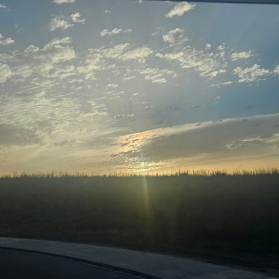 The image depicts a landscape scene, possibly at sunrise or sunset, with a focus on the sky and a silhouetted horizon. The sky is filled with a multitude of small, scattered clouds that appear textured and layered, with some areas of brighter illumination suggesting the sun's position. The horizon is marked by a dark, silhouetted line of vegetation or terrain, with sparse vertical elements suggesting dried grasses or thin trees. A faint beam of light or sun flare emanates from the lower center of the image, illuminating the lower sky and casting a glow on the horizon. In the foreground, a portion of a paved surface, likely a road or sidewalk, is visible, suggesting an urban or semi-urban setting. No people or specific landmarks are identifiable. The image does not provide any discernible features that would allow for definitive identification of the location as Illescas, Spain.