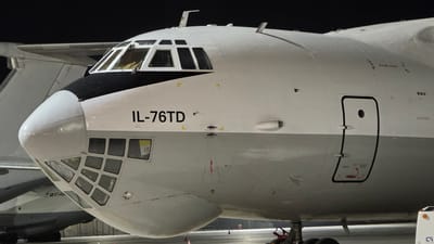 This is a close-up, exterior, eye-level, medium shot of the front section of an IL-76TD aircraft parked on a tarmac at night. The aircraft is predominantly white with a black nose section and grey lower fuselage. The cockpit windows are visible at the top, and below them is the distinctive glazed nose section of the aircraft. The text "IL-76TD" is clearly visible in black lettering on the white fuselage. A closed passenger door with a small circular window is visible on the right side of the image. The scene is illuminated by artificial lighting, as indicated by the dark background. The ground beneath the aircraft appears to be concrete, and some orange traffic cones are visible in the lower portion of the image. No people or actions are depicted. The image focuses solely on the static presentation of the aircraft.