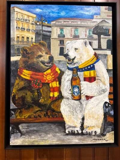 The image is a painting depicting two anthropomorphic bears sitting on a park bench in a city. The bear on the left is a brown bear wearing a scarf with a sun and flowers. The bear on the right is a polar bear wearing a scarf and holding a bottle of "POLAR" beer. In the background, there are buildings and a blue sky with clouds. The painting is signed "MAVANS, '20" in the lower right corner. There is no information in the image that suggests it is located in Torrejón de Ardoz, Spain.