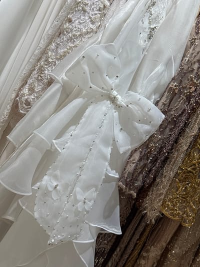 The image displays a close-up view of several ornate dresses, likely wedding or formal gowns, hanging side-by-side. The dominant dress in the foreground is white with a large, decorative bow adorned with pearls and sequins. This dress features tiered ruffles and delicate floral appliqués, also embellished with pearls and beads. To the right of the white dress are other gowns in muted tones of brown, taupe, and gold, showcasing intricate beadwork, sequins, and lace. The background is out of focus, suggesting the dresses are in a retail setting such as a boutique or bridal shop. There are no people or discernible environmental features beyond the dresses themselves. The scene is static and well-lit, emphasizing the texture and detail of the garments. This image was taken in the city of Tanjungmeru, Indonesia.