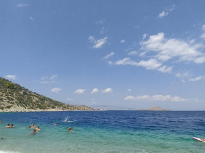 An aerial view shows a beach with people swimming and a distant island in the Municipal Unit of Solygeia, Greece. The water is clear and blue, with a rocky coastline in the background. Several people are visible in the water, some swimming and others relaxing. A small island is located in the distance, and clouds are scattered across the sky. The scene is calm and serene, suggesting a pleasant day at the beach.
